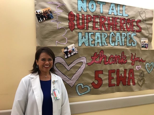 Female health care manager wearing a white lab coat, standing in front of a banner that says, "Not all superheros wear capes."